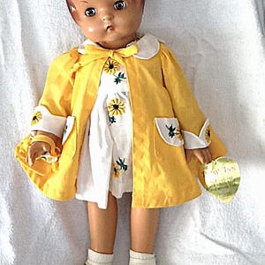 May include: Vintage doll wearing a yellow coat with white trim and pockets, over a white dress with embroidered yellow flowers. The doll has brown hair, a serious expression, and is wearing white shoes. A small yellow purse is in the doll's hand.