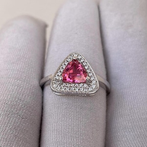 May include: A silver ring with a triangular, bright pink gemstone. The gemstone is encircled by a halo of small, clear accent stones. The ring's band is a simple, polished silver.