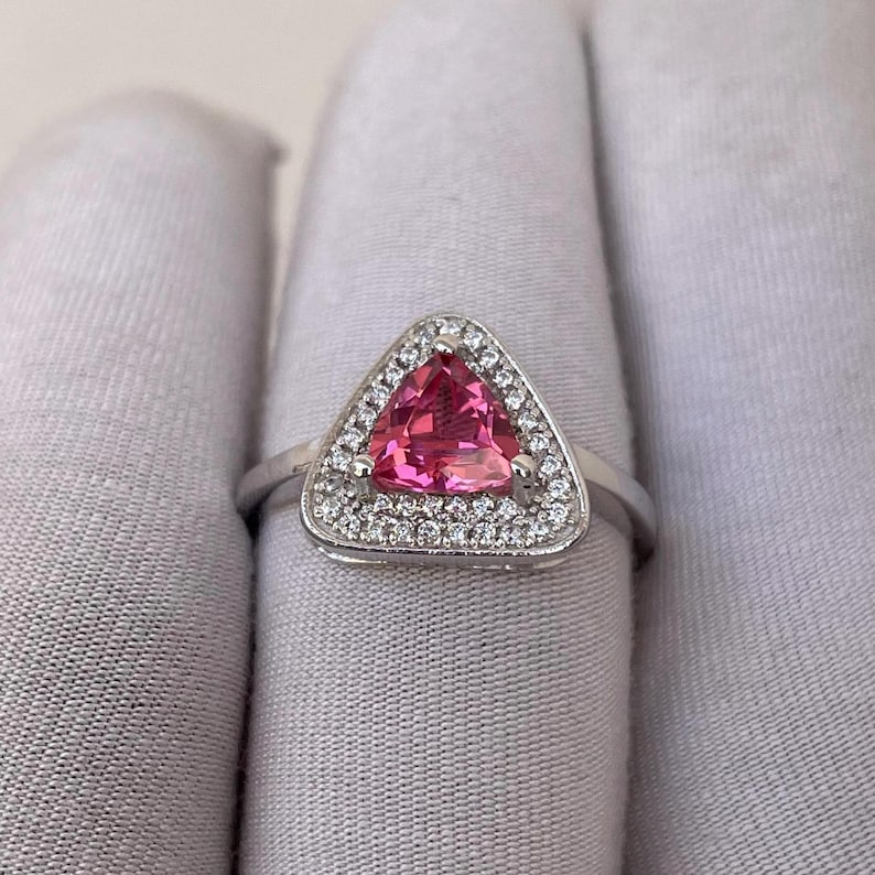 May include: A silver ring with a triangular, bright pink gemstone. The gemstone is encircled by a halo of small, clear accent stones. The ring's band is a simple, polished silver.