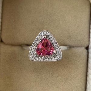 May include: A silver ring featuring a triangular, vibrant pink gemstone, surrounded by a halo of small, clear gemstones. The ring is set against a neutral background, highlighting the sparkle and color of the jewelry.
