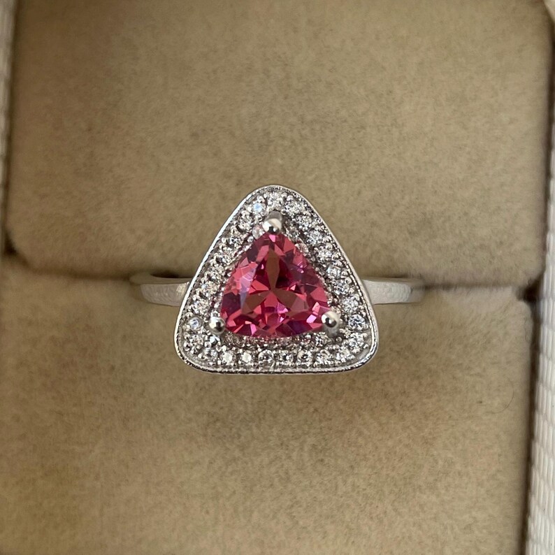 May include: A silver ring with a triangular, vivid pink gemstone, encircled by a halo of small, clear gemstones. The ring is displayed against a neutral backdrop, emphasising the sparkle and colour of the jewellery.