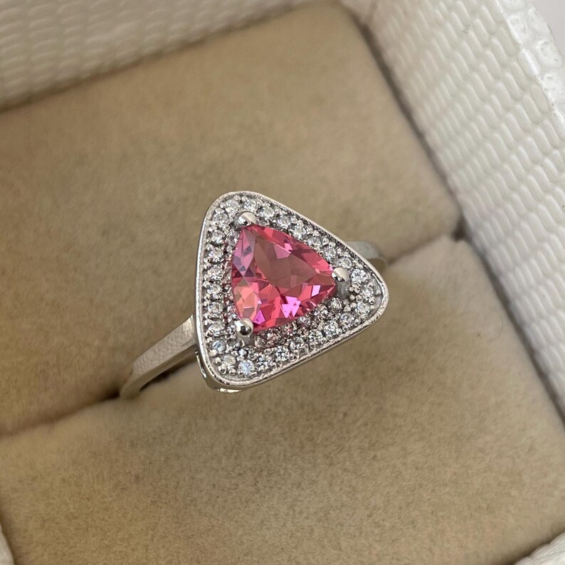 May include: A silver ring with a triangular pink gemstone at its centre, encircled by small, clear stones. The ring has a slender, polished band and a detailed setting. The ring is presented in a neutral-toned jewellery box.