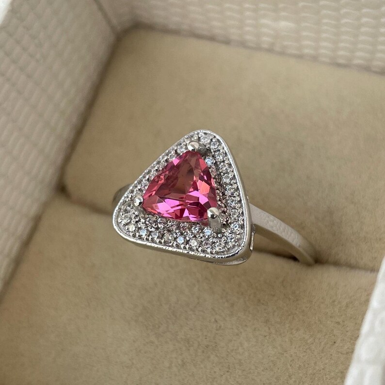 May include: A silver ring with a triangular, pink gemstone at its centre, encircled by a halo of small, clear stones. The ring has a silver band, and the design is elegant and striking.