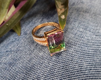 May include: A gold ring with a large, rectangular, multi-colored gemstone. The gemstone has shades of pink, green, and purple.