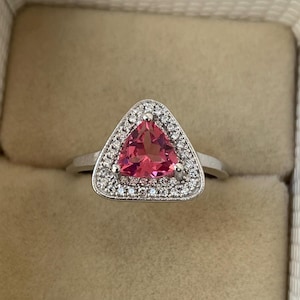 May include: A silver ring with a triangular, bright pink gemstone, encircled by a halo of small, clear stones. The ring is set against a neutral backdrop, emphasising the sparkle of the gemstones.