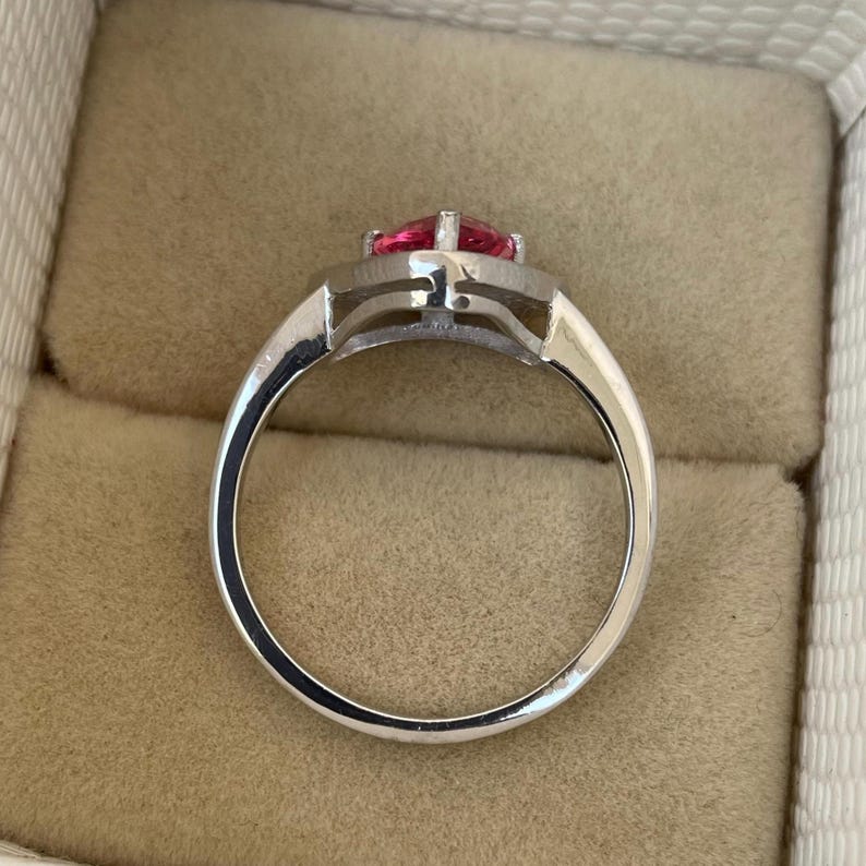 May include: A silver ring featuring a heart-shaped, bright pink gemstone. The ring has a modern design, with a polished band and a detailed setting. The ring is presented in a jewellery box. The gemstone is a vibrant shade of pink.
