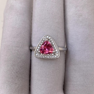 May include: A silver ring with a triangular, bright pink gemstone, encircled by a halo of small, clear gemstones. The ring's slender, polished band adds to its elegant design. The ring is set against a neutral background.