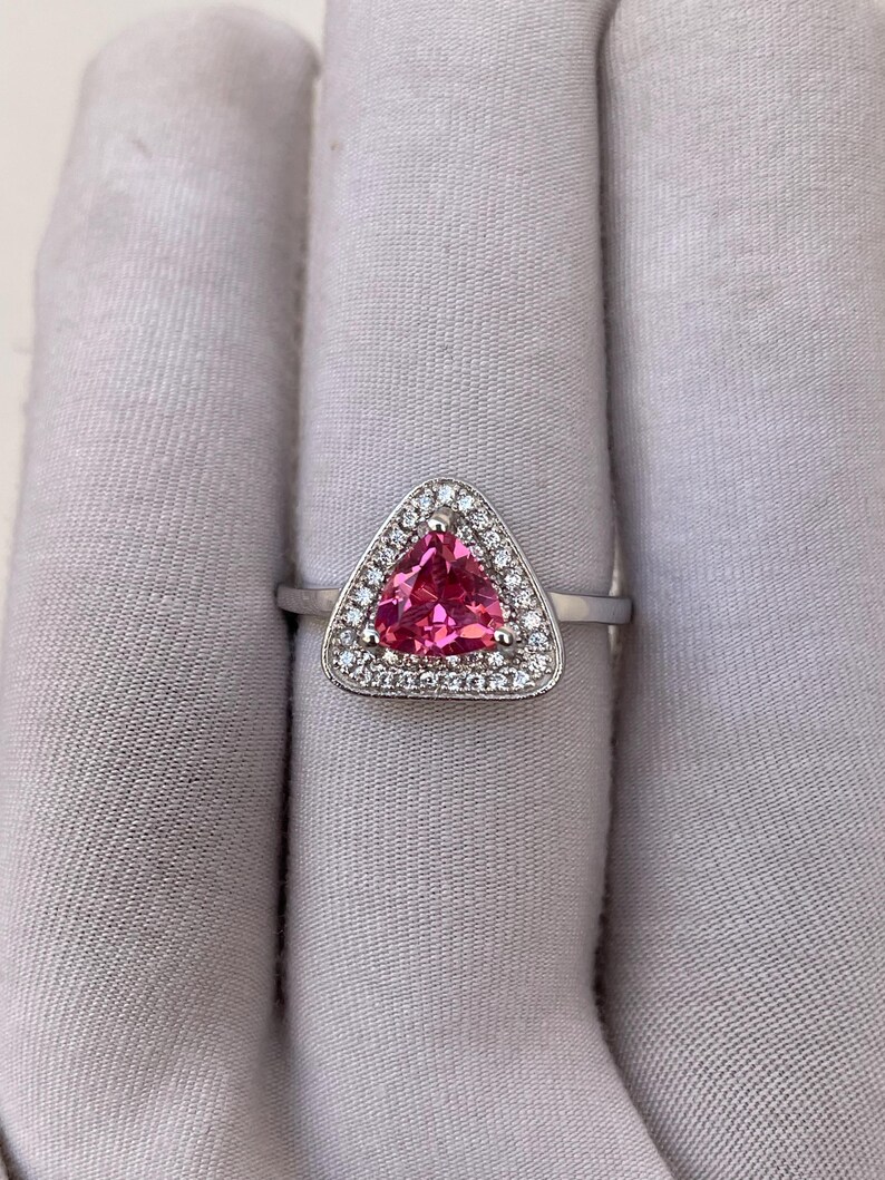 May include: A silver ring with a triangular, bright pink gemstone, encircled by a halo of small, clear gemstones. The ring's slender, polished band adds to its elegant design. The ring is set against a neutral background.