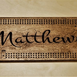 May include: A wooden cribbage board with a dark brown stain. The board has a black engraved border with holes for pegs. The name "Matthews" is engraved in the centre of the board.