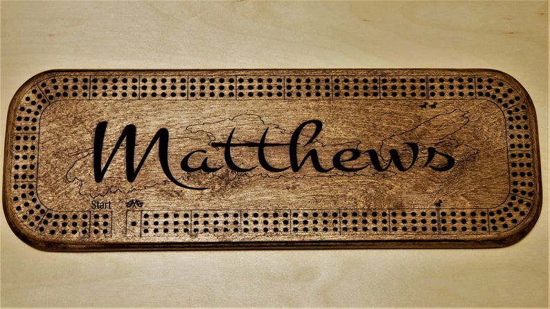 May include: A wooden cribbage board with a dark brown stain. The board has a black engraved border with holes for pegs. The name "Matthews" is engraved in the centre of the board.