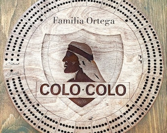 May include: A wooden cribbage board with a brown stain and a whitewash finish. The board features a shield with a profile of a person wearing a head covering. The text "Familia Ortega" and "COLO-COLO" are engraved on the board.