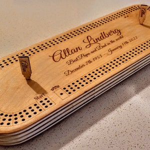 May include: A wooden cribbage board with a personalised inscription that reads "Allan Lindberg Best Papa and Dad in the world December 7th 1953 - January 11th 2022". The board has a start position and two pegs.