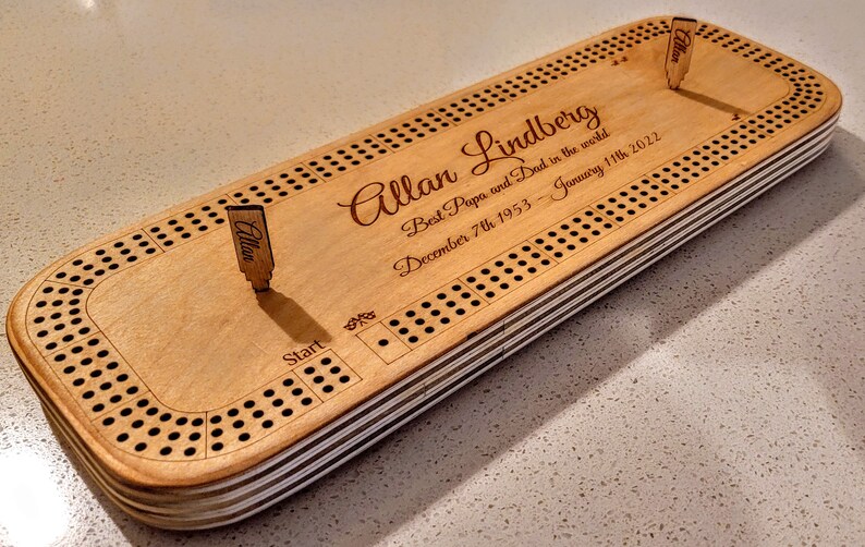 May include: A wooden cribbage board with a personalised inscription that reads "Allan Lindberg Best Papa and Dad in the world December 7th 1953 - January 11th 2022". The board has a start position and two pegs.