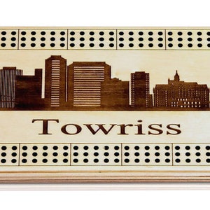 May include: A wooden cribbage board featuring a skyline of buildings and a bridge. The board is engraved with the word "Towriss".