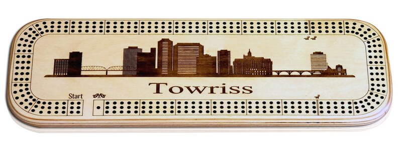 May include: A wooden cribbage board featuring a skyline of buildings and a bridge. The board is engraved with the word "Towriss".