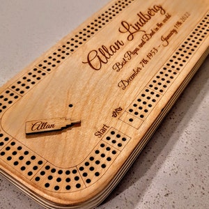 May include: A wooden cribbage board with a personalised inscription for "Allan Lindberg" and the dates "December 7th 1953 - January 11th 2022". The inscription reads "Best Papa and Dad in the world".