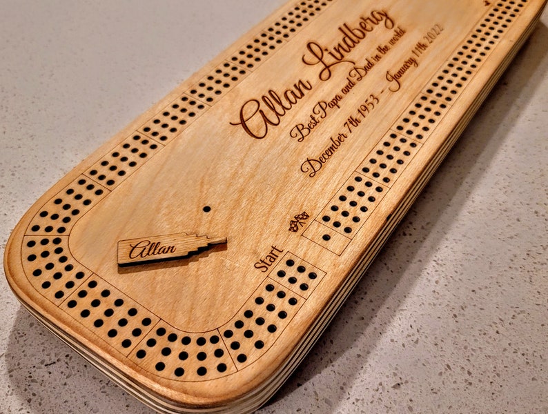 May include: A wooden cribbage board with a personalised inscription for "Allan Lindberg" and the dates "December 7th 1953 - January 11th 2022". The inscription reads "Best Papa and Dad in the world".
