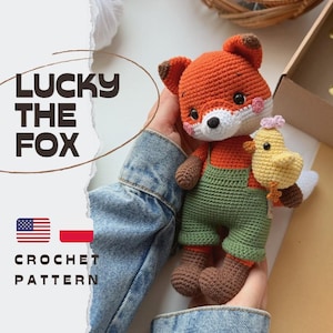 Op de afbeelding: Een gehaakt vossenspeeltje van oranje, wit en bruin garen, met een groene tuinbroek aan en een klein geel kuikentje in de hand. De afbeelding bevat de tekst LUCKY THE FOX en CROCHET PATTERN, samen met Amerikaanse en Poolse vlagiconen.