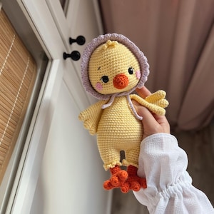May include: A crocheted yellow chick toy with a purple hooded hat and orange feet. The chick has black eyes and a small orange beak.
