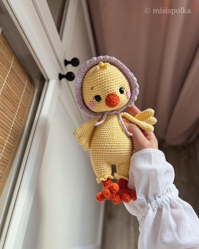 May include: A crocheted yellow chick toy with a purple hooded hat and orange feet. The chick has black eyes and a small orange beak.