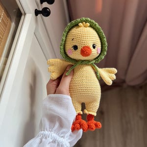 May include: A crocheted yellow chick toy with a green hooded hat and orange feet. The chick has black eyes, a pink blush, and an orange beak.