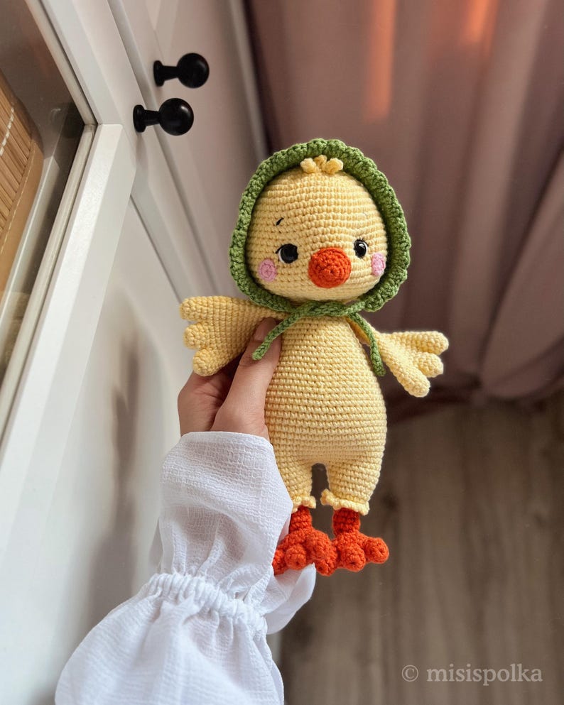 May include: A crocheted yellow chick toy with a green hooded hat and orange feet. The chick has black eyes, a pink blush, and an orange beak.