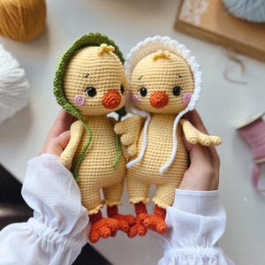 May include: Two crocheted yellow chicks with orange beaks and feet. One chick is wearing a green hooded hat and the other is wearing a white hooded hat.