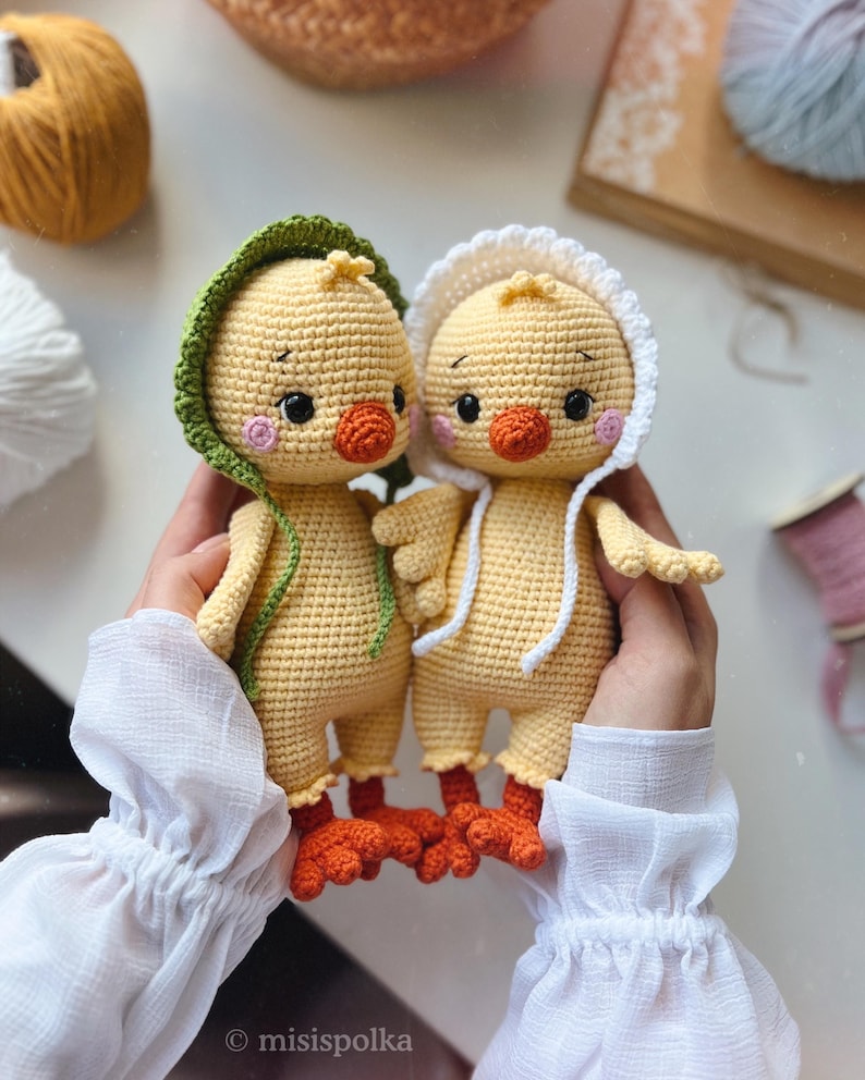 May include: Two crocheted yellow chicks with orange beaks and feet. One chick is wearing a green hooded hat and the other is wearing a white hooded hat.