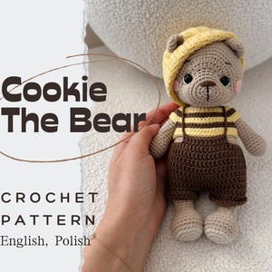 Op de afbeelding: Een gehaakte berenpop genaamd "Cookie The Bear" met een gele hoed, een gestreept shirt en een bruine overall. De pop heeft een lichtbruin lichaam, roze wangen en zwarte ogen. De afbeelding bevat ook de tekst "CROCHET PATTERN English, Polish."