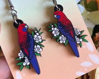 May include: A pair of earrings featuring a red and blue bird with white flowers on a wooden background. The earrings are made by Hyper Finch Enamel Pins and Accessories.