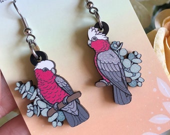 May include: A pair of earrings featuring pink and grey cockatoos with green foliage, on a card that reads "HYPER FINCH Enamel Pins and Accessories".
