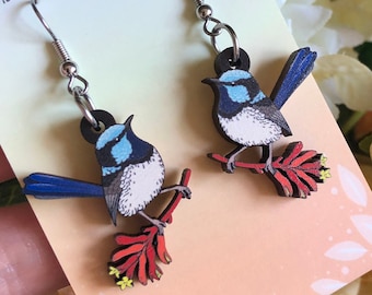 May include: A pair of dangle earrings featuring a blue and white bird perched on a red and yellow floral branch. The earrings are made of wood and have silver-colored hooks. The card behind the earrings reads "HYPER FINCH Enamel Pins and Accessories."