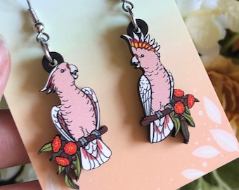 May include: A pair of earrings featuring pink cockatoos with red berries on a branch. The earrings are made of wood and have silver hooks. The earrings are displayed on a card that says "HYPER FINCH Enamel Pins and Accessories".