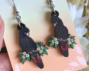 May include: A pair of black cockatoo earrings with a detailed design. Each earring features a black bird perched on a branch with white flowers and green leaves. The earrings are hanging from silver-colored hooks. The text "HYPER FINCH Enamel Pins and Accessories" is visible.
