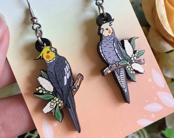 May include: A pair of earrings featuring a hand-painted wooden design of two cockatiels perched on branches with white flowers. The earrings are silver and have a hook closure. The earrings are displayed on a card with the text "HYPER FINCH Enamel Pins and Accessories".