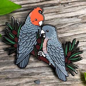 Könnte beinhalten: Zwei graue und rote Kakadus mit schwarzen Augen, die auf einem Ast mit grünen Blättern und roten Beeren sitzen. Die Vögel schauen sich an und haben ein detailliertes Design.