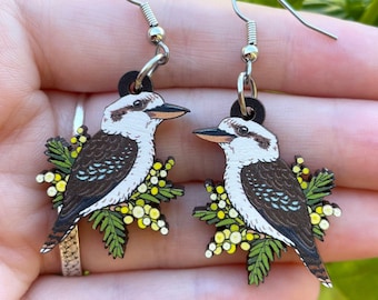 May include: A pair of wooden earrings featuring a detailed illustration of a kookaburra bird. The bird has a white head, brown wings, and is perched on a sprig of yellow flowers and green leaves. The earrings have silver-colored hooks.