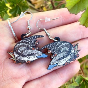 May include: A pair of dangle earrings with blue-tongued lizard designs. The earrings have silver hooks and show the lizards with grey and white scales, blue tongues, and small red accents. A unique accessory.