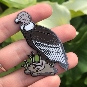 May include: An enamel pin depicting an Andean condor, a large bird of prey, perched on a rock with green grass. The condor has a white neck ruff and a brown body with white markings on its wings.