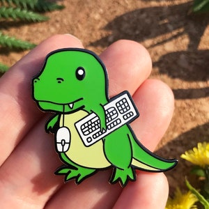 Peut inclure: Épinglette en émail de dinosaure vert avec un clavier blanc et une souris blanche. Le dinosaure porte une chemise jaune.