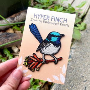 May include: An embroidered iron-on patch depicting a vibrant bird with blue, turquoise, black, and white feathers, perched on a red branch. The patch is on a peach-coloured card with the text "HYPER FINCH" and "Iron-on Embroidered Patch".
