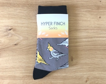 May include: A pair of gray socks with a colorful bird pattern. The socks have a black cuff and a white tag that says "HYPER FINCH Socks".