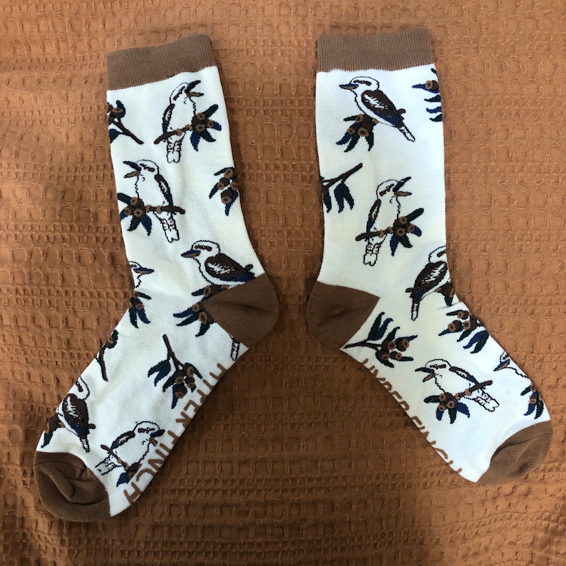 May include: A pair of white socks with a brown cuff. The socks feature a repeating pattern of kookaburras, a type of bird native to Australia.