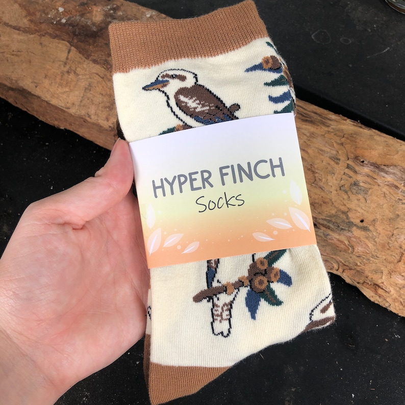 May include: A pair of white socks with a brown cuff. The socks feature a colorful print of kookaburras and eucalyptus leaves. The socks are packaged with a label that reads "HYPER FINCH Socks".