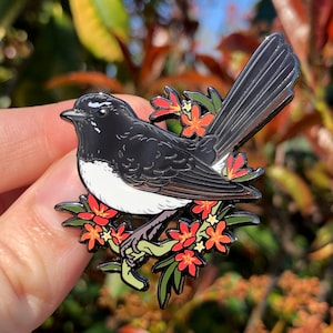May include: A decorative bird pin featuring a black and white bird design. The bird is set against a backdrop of red flowers and green leaves. The pin is made of metal and has a polished finish.