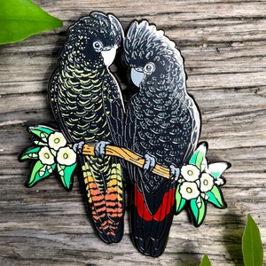 Op de afbeelding: Zwarte en gele emaille pin met twee zwarte kaketoes die op een tak zitten met witte bloemen. De kaketoes kijken elkaar aan en hebben zwarte veren met gele accenten.