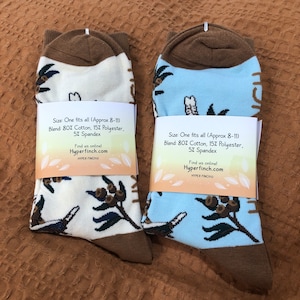 May include: Two pairs of socks with a brown cuff and a white and blue background. The socks feature a repeating pattern of birds and leaves. The socks have a tag that reads "Size: One fits all (Approx 8-11) Blend: 80% Cotton, 15% Polyester, 5% Spandex Find us online! Hyperfinch.com HYPER FINCHO"