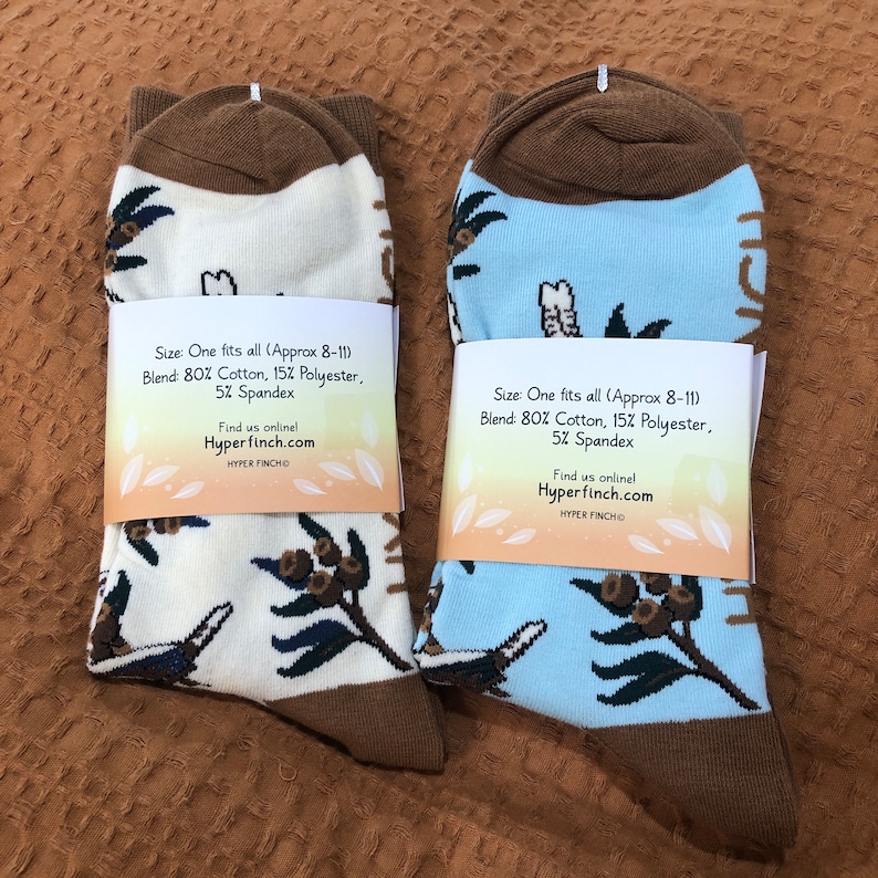 May include: Two pairs of socks with a brown cuff and a white and blue background. The socks feature a repeating pattern of birds and leaves. The socks have a tag that reads "Size: One fits all (Approx 8-11) Blend: 80% Cotton, 15% Polyester, 5% Spandex Find us online! Hyperfinch.com HYPER FINCHO"