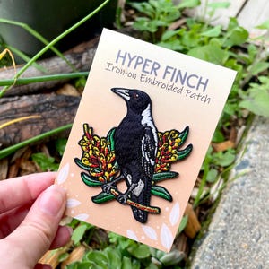 May include: An embroidered patch of a black and white bird on a branch with yellow and red flowers. The patch is on a peach card with the text "HYPER FINCH" and "Iron-on Embroidered Patch". Ideal for clothing or accessories.