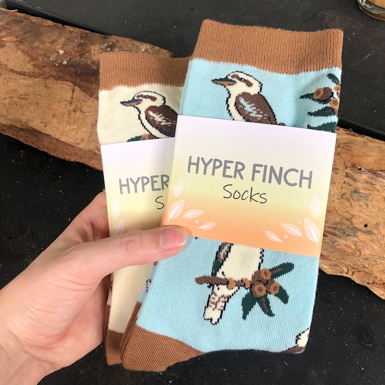 May include: Two pairs of socks with a brown cuff and a blue background. The socks feature a cartoon image of a kookaburra bird. The socks are packaged with a tag that reads "HYPER FINCH Socks".